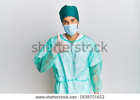 Young handsome man wearing surgeon uniform and medical mask doing happy thumbs up gesture with hand. approving expression looking at the camera showing success. 