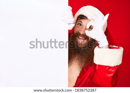 Handsome young red head man with long beard wearing santa claus costume holding banner smiling happy doing ok sign with hand on eye looking through fingers 
