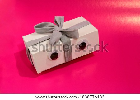 Funny white gift box with a bow and plastic eyes on a pink background. Blank with copy space.