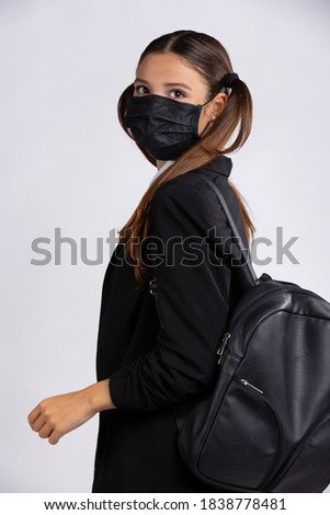 A young female student wearing a black face mask to prevent infection with the corona virus, wears a black jacket and a backpack. Photo on gray background