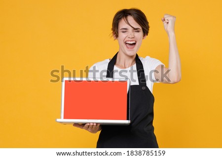 Joyful young woman 20s barista bartender barman employee in apron hold laptop computer with blank empty screen mock up copy space doing winner gesture isolated on yellow background studio portrait