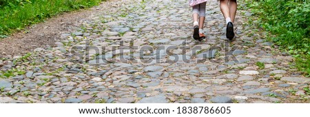 Legs of young man and woman walking along old rural paved road with large rocks between lush green grass on sides on nice spring day close backside view.