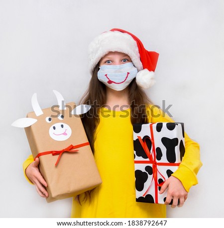 crazy exited happy girl with gifts in the red christmas hat and medical mask on the white background. wrapping paper with cow spots.