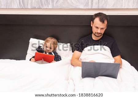 Caucasian father is working from home with laptop in bed and kid is near, watching cartoons, white linen, isolated period at lockdown with covid, remote work concept.
