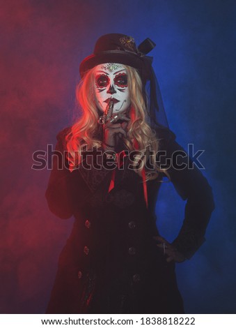Beautiful woman with scary Halloween make up dead day calavera style with finger on lips on blue and red smoke background

