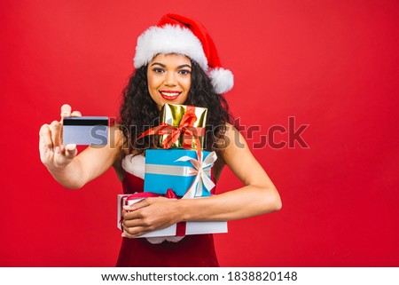 Beautiful portrait of young african american woman happy holding credit card and gift box in Christmas holiday isolated on red background, surprised girl shopping with credit card in xmas day.