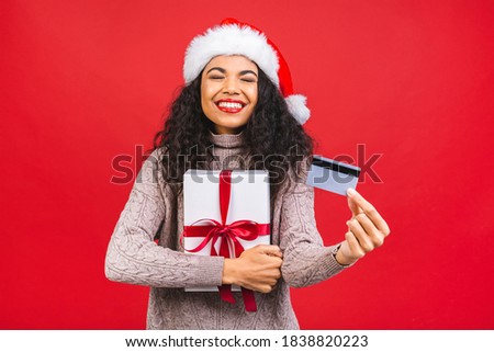 Beautiful portrait of young african american woman happy holding credit card and gift box in Christmas holiday isolated on red background, surprised girl shopping with credit card in xmas day.