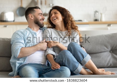 Happy married young couple hugging, sitting on cozy couch together, overjoyed laughing woman and man having fun, enjoying leisure time, relaxing on sofa in living room at home, good relationship Royalty-Free Stock Photo #1838823604