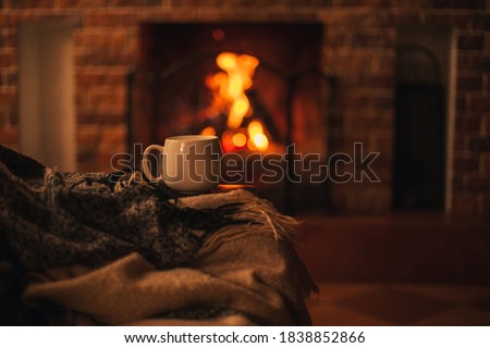 Mug with hot tea standing on a chair with woolen blanket in a cozy living room with fireplace. Cozy winter day.