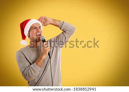 Emotional man in Santa Claus hat singing with microphone on yellow background, space for text. Christmas music