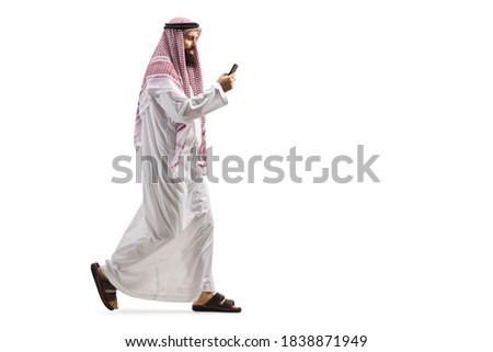 Full length profile shot of an arab man using a smartphone and walking isolated on white background