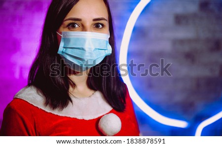 Young woman in medical mask and Santa Claus suit against illuminated wall. Brunette in Christmas costume and protective mask. Concept of safe Christmas celebration during coronavirus pandemic.