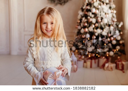 Cute little blonde girl smiles holds a gift box next to the Christmas tree. Christmas morning concept, space for text