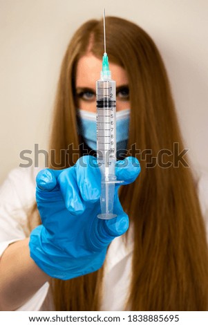 A hand of a young nurse with protective mask face and blue gloves holding a syringe close-up