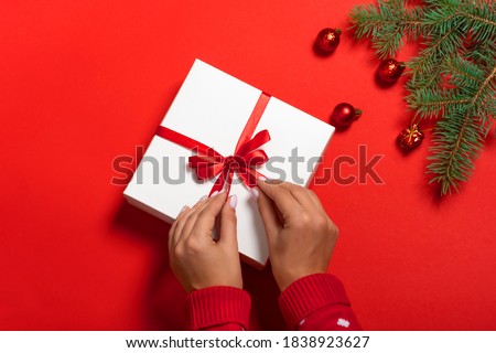 Female hands in a red new year sweater holding a white gift box on a red background, fir tree, christmas baubles, toys with copy space. Flat lay. Festive background.