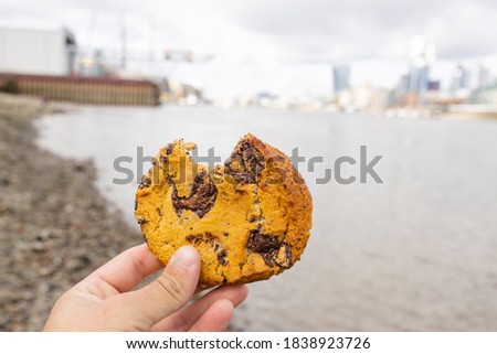 Landscape View of a Hand Holding a Bitten Chocolate Chips Cookie with a Blurry River Thames and CIty as Background