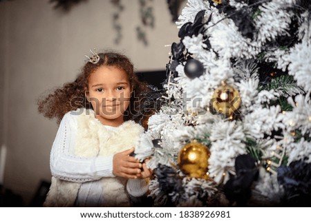 Beautiful mixed little girl  waiting for a miracle on the background of Christmas trees in dark interior of the house.
