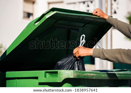Man throwing out black eco-friendly recyclable trash bag in to big plastic green garbage container. Take out the trash Royalty-Free Stock Photo #1838957641