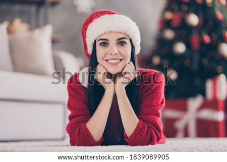 Photo of charming young girl lay carpet fists chin toothy smiling wear santa headwear red pullover in decorated living room indoors