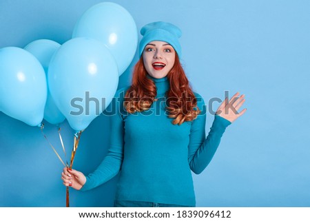 Image of happy young woman celebrating birthday with air balloons, looking at camera and raising hand, wearing casual shirt and cap, has red curls and red lips.