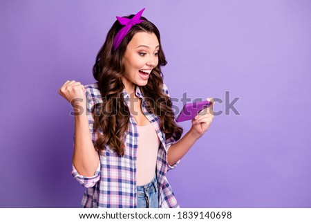 Photo of delighted girl raise fists win game on cellphone wear checkered shirt isolated over purple color background