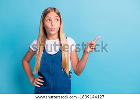 Photo of surprised teenager blond long girl point look empty space wear white t-shirt denim dress isolated on blue color background