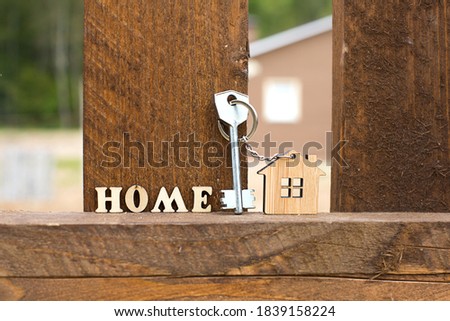 Keychain of wooden figure house on fence with key and inscription in English letters "home". Cottage in the background. Building, project, moving to new house, mortgage, rent and purchase real estate.
