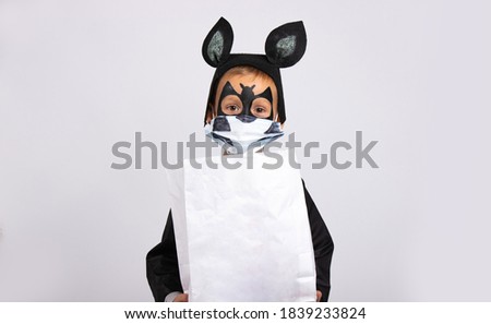  Halloween, little cute boy dressed as a bat holding a white bag of chocolates with empty space .. Trick or treat. Child in a medical mask on a gray background. The virus has attacked the world