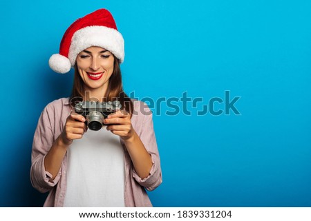 Smiling young woman in shirt wearing santa claus hat holding looking at retro camera on blue background.