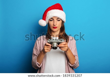 Young woman in santa claus hat looks thoughtfully at a retro camera on a blue background.