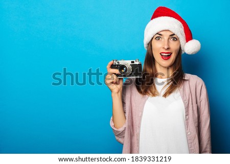 Surprised young woman posing on blue background wearing santa claus hat holding retro camera.
