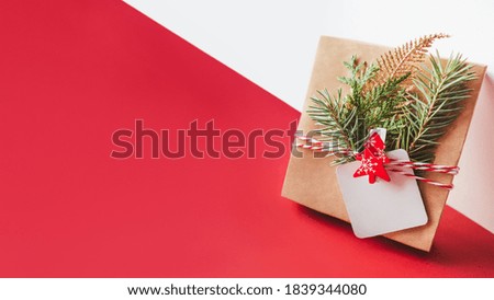 New Year present on geometrical white and red festive background. Gift wrapped in craft paper with fir tree twigs, red Christmas tree and clear tag. Copy space on label. Winter holiday spirit.