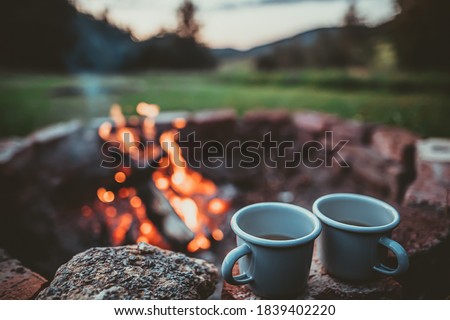 Campsite With Fire Pit and Two Tin Cups with hot tea. Burning Campfire with mountain landscape with evening sunset sky over the forest and hills. Royalty-Free Stock Photo #1839402220