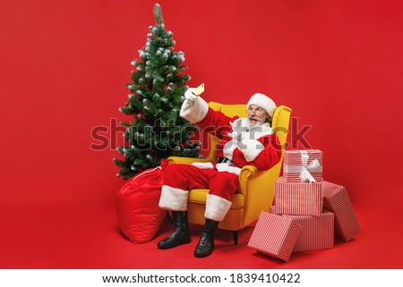 Excited Santa Claus man in Christmas hat suit sit in armchair with fir tree gifts doing selfie shot pointing on mobile phone isolated on red background. Happy New Year celebration holiday concept