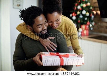 Happy black woman embracing her husband and giving his a gift on Christmas day at home. 
