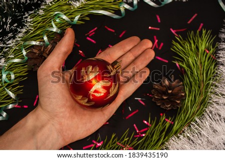 Holding patterned Christmas ball toy in hand on black background with red confetti scattered among pine branches and cones with colored curly ribbons. Decorating Christmas tree concept