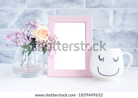 mockup photo frame, white mug with a smiling face and closed eyes and a vase with a rose and gypsophila.