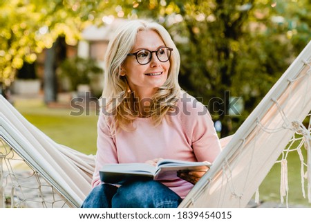 Smiling senior good-looking blond woman wearing glasses while reading in hammock in the summer garden Royalty-Free Stock Photo #1839454015