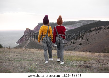 Back view of two preteen girls traveler wearing denim overalls, yellow and red sweater and knitted hats sit on top of the mountain landscape and looking to the sea. 