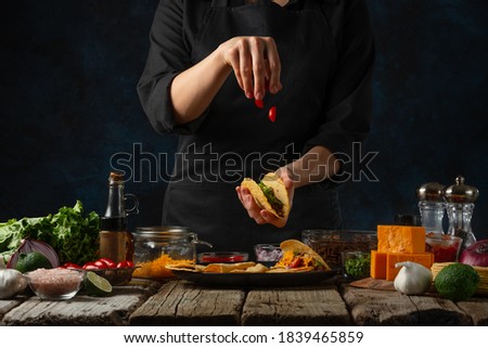 Professional chef in black uniform puts chopped tomato on corn tortilla for cooking tacos on dark blue background. Traditional mexican cuisine. Concept of tasty street food. Frozen motion.