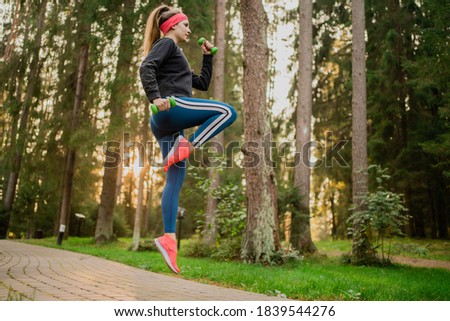 enhanced fitness training in a Park in the city center, doing jumps on the spot exercises with dumbbells in her hands, confident athletic woman in comfortable fashionable sportswear