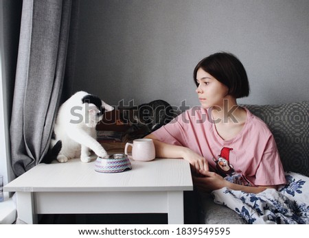 A young woman with short hair dines with her cats. Pets eat from a bowl. The cat is on the table. Love to the animals. The concept of living with a pet. 