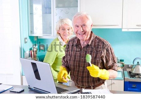 Horizontal waist up portrait of a joyous senior couple using a laptop and man doing household chores looking at the camera.