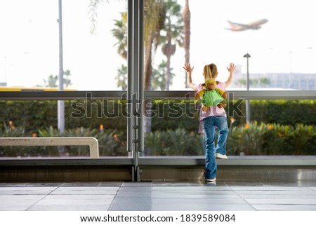 A wide rear view shot of a girl with a monkey rucksack leaning and looking through a large window in an airport lounge watching an airplane taking off.