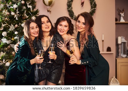 Portrait of four beautiful women in evening dresses who celebrate the new year or Christmas in a beautiful interior with a dressed Christmas tree, garlands and a drink in hand