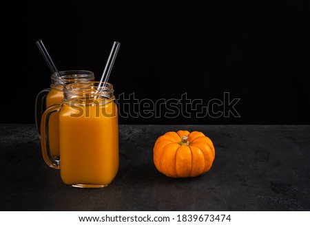 Two square glass jars with non-alcoholic pumpkin mocktail and glass straws on black textured surface. Whole pumpkin is near jars. Horizontal background for Halloween with copy space. Selective focus.