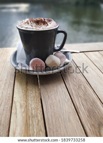 Grey mug of hot chocolate topped with whipped cream, chocolate powder and marshmallows on the side sitting on a wooden table with water in the background. Close-up plan.