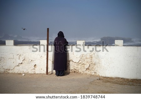 Man with dark Gandora short sleeved robe looking at wild sea before storm, Essaouira, Morocco