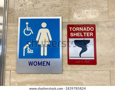 The women's restroom and tornado shelter signs directing people to the shelter in a public building.