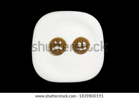 Two ginger cookies with happy and sad face on a white plate on a black background.